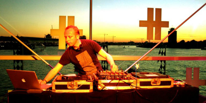 Sound artist Scanner djing at sunset in a building with large windows. There is water and a city scape in the background.